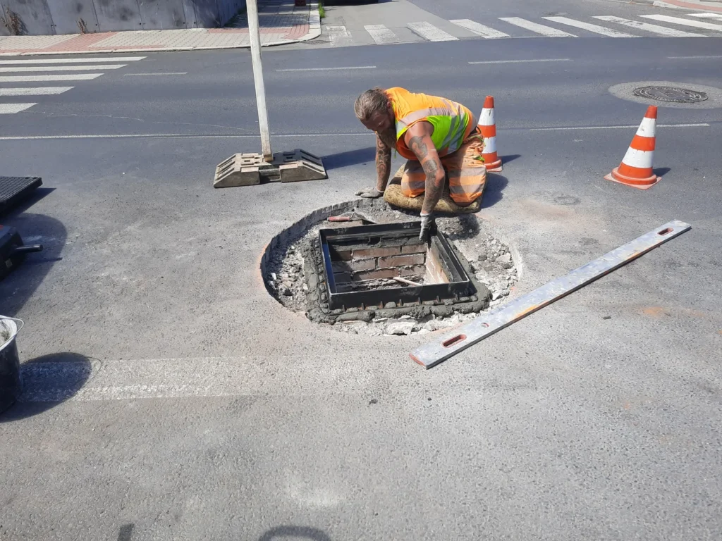 Dělník osazuje čtvercový rám kanalizačního poklopu na maltové lože při zabudování, vedle otevřená šachta a dopravní kužely